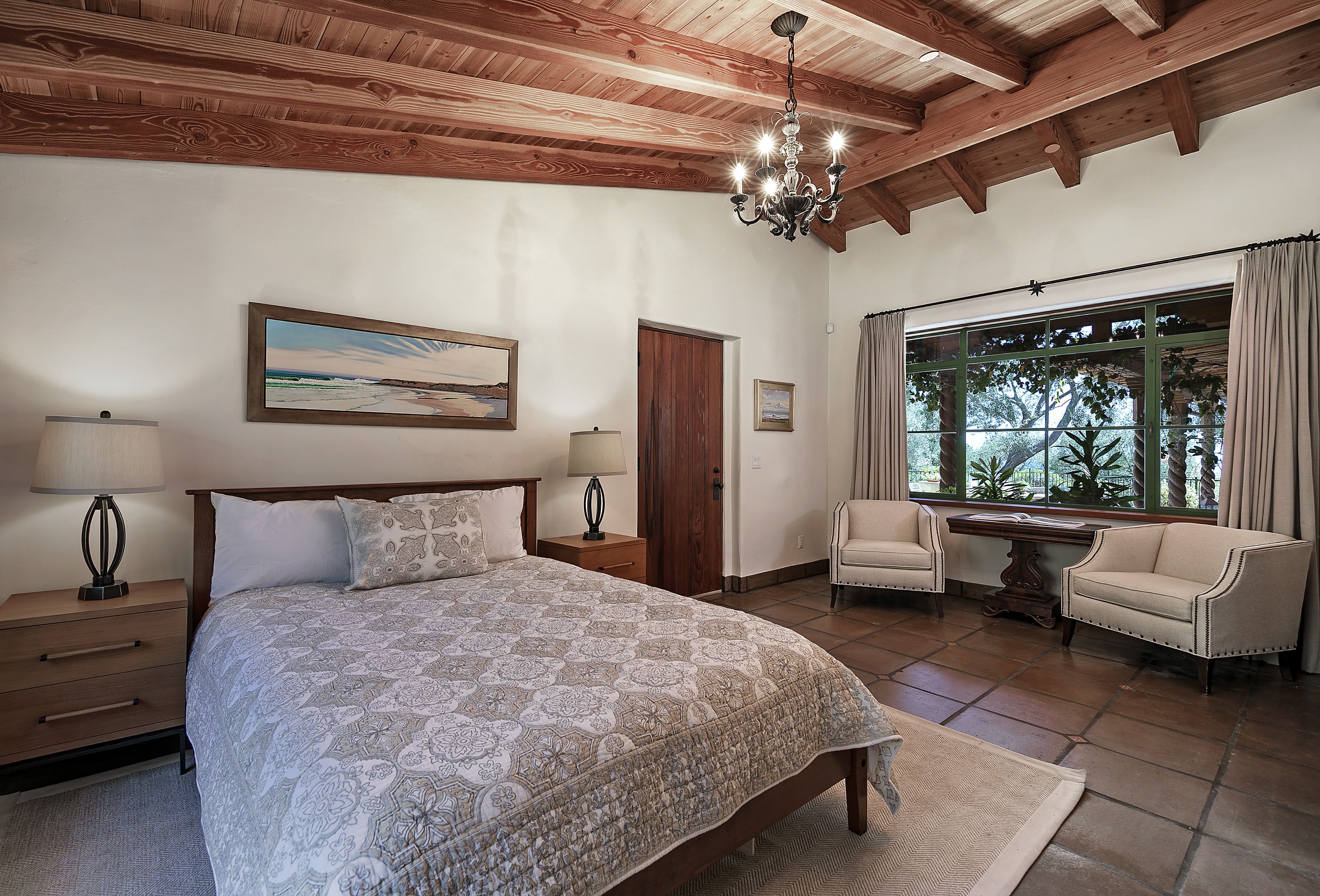 Furnished bedroom with open-beam ceiling, and large window with view of vine-covered pergola and trees
